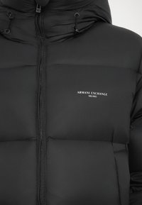 Sort pufferjakke med lynlås foran, høj krave og justerbare snore; har quiltet tekstur og et hvidt logo på brystet.