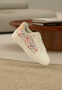 Λευκό αθλητικό παπούτσι με ροζ και μοβ λουλουδάτα σχέδια, μπεζ τακούνι, λευκά κορδόνια, τοποθετημένο σε μοντέρνο καθιστικό.