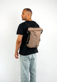 Barna roll-top hátizsák, szürke rugalmas zsinór díszítéssel, fekete pántokkal és matt felülettel. A felhasználó fekete pólót és világos farmernadrágot visel.