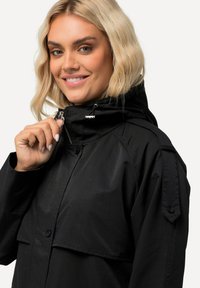 Musta takki, jossa on korkea kaulus, vetoketjusuljenta ja nappikoristeet. Takissa on tekstuurinen kangas ja etupuolella tasku.