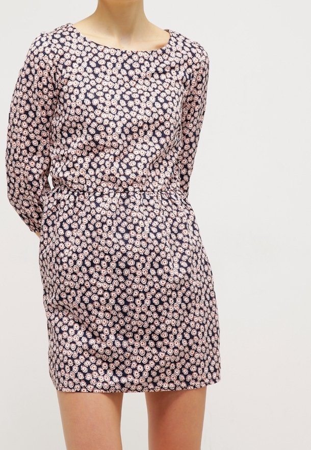 Kobieta w dopasowanej, długiej sukience z granatowym tłem i małym, jasnoróżowym wzorem kwiatowym, stojąca na jednolitym tle.