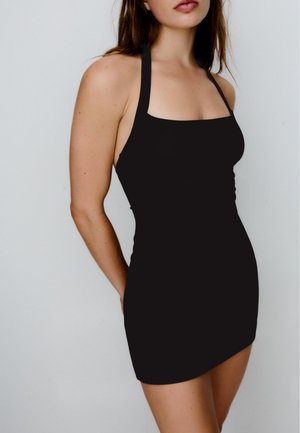 Junge Frau trägt ein ärmelloses schwarzes Neckholder-Minikleid und steht vor einem schlichten hellen Hintergrund.