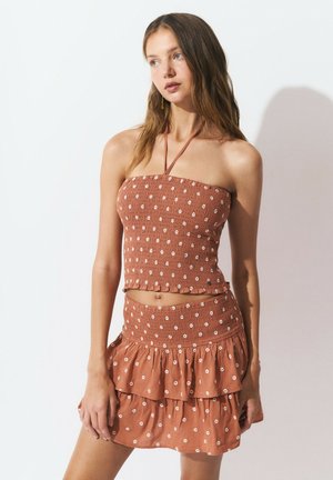 Top corto marrón con fruncido y patrones florales blancos, combinado con una falda a juego con volantes. Diseño de cuello halter, textura suave y bordes con volantes.