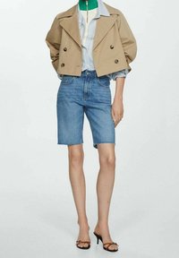 Modella che indossa un giacca doppiopetto beige cropped su una camicia a righe, pantaloni corti in denim alla ginocchia e sandali con tacco marroni su uno sfondo neutro.