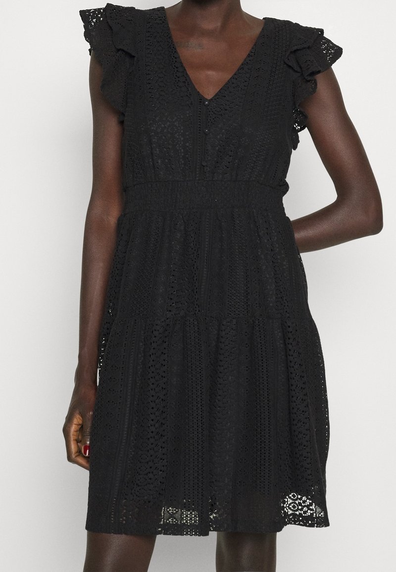Robe en dentelle noire avec des manches volantées, un col en V et une taille élastique portée par une personne à la peau foncée sur un fond clair uni.