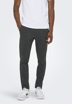 Mand iført mørkegrå bukser med striber, hvide sneakers med grønt logo og en hvid kortærmet skjorte, hænder afslappet langs siden.