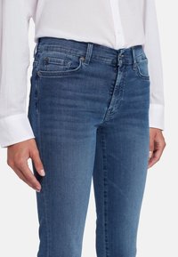 Jeans in denim blu scuro con una vestibilità slim, dotati di cinque tasche e dettagli in bottone metallico. Abbinati a una camicia bianca a maniche lunghe.