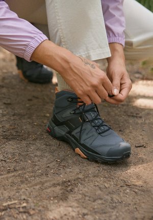 Stivali da trekking grigio scuro con dettagli arancioni, sistema di allacciatura, materiale testurizzato e suole robusti, posati su terra con una mano che stringe i lacci.