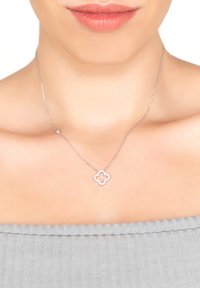 Silver necklace featuring a clover-shaped pendant with a cut-out center, worn on bare skin. The background is a textured gray garment.