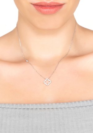 Silver necklace featuring a clover-shaped pendant with a cut-out center, worn on bare skin. The background is a textured gray garment.