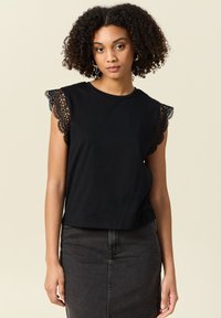 Svart bomullströja med spetsdetaljer på korta ärmar, rund halsringning och en avslappnad passform. Bärs med en mörk denimkjol.