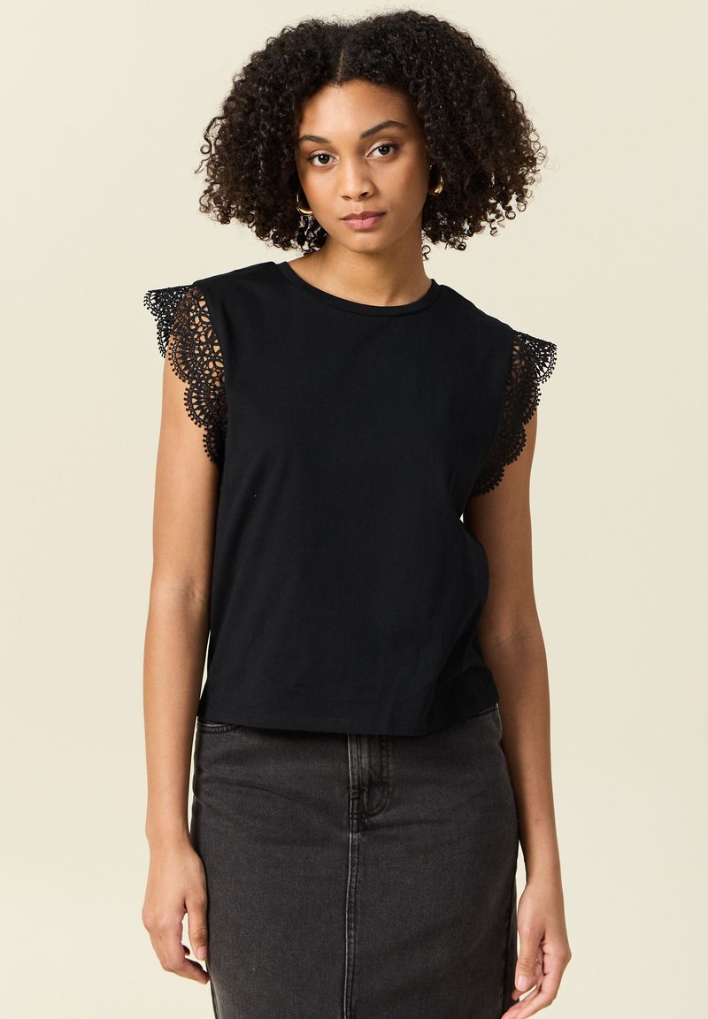 Svart bomullströja med spetsdetaljer på korta ärmar, rund halsringning och en avslappnad passform. Bärs med en mörk denimkjol.