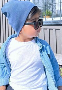 Lapsi, joka on pukeutunut vaaleansiniseen ribattuun pipoon ja tummiin aurinkolaseihin, valkoisen paidan ja nappikiinnitteisen farkkutakin päälle.