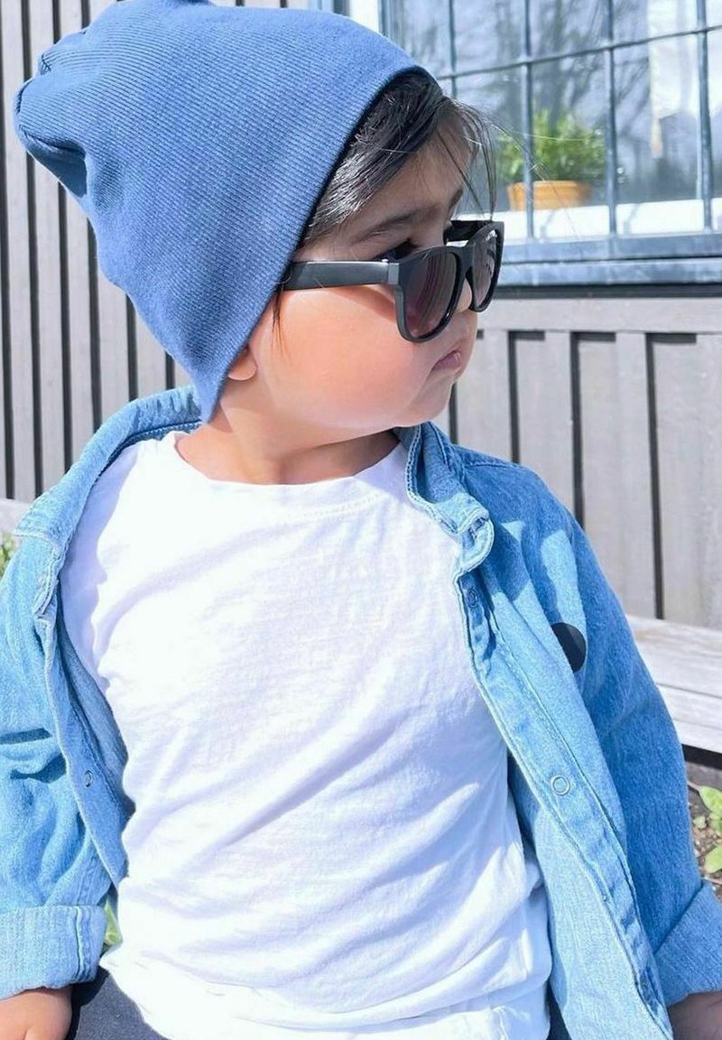 Lapsi, joka on pukeutunut vaaleansiniseen ribattuun pipoon ja tummiin aurinkolaseihin, valkoisen paidan ja nappikiinnitteisen farkkutakin päälle.