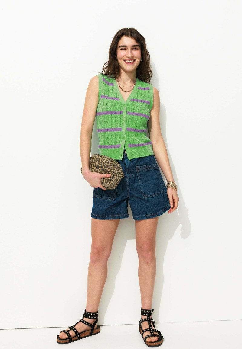 Gilet en tricot vert avec des rayures violettes, shorts en denim, pochette à imprimé animal, et sandales noires à lanières. Différentes textures et motifs visibles.