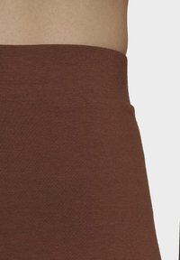 Detailní záběr na pas osoby, která má na sobě hnědý, žebrovaný kus oděvu s vysokým pasem, ukazující část odkryté pokožky nad pasem.