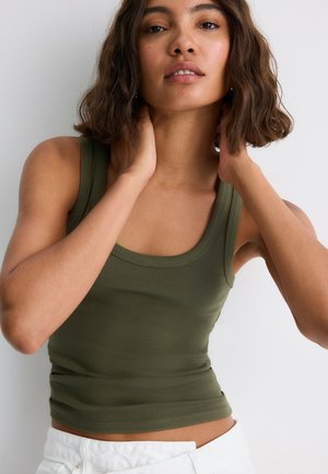 Femme aux cheveux foncés et ondulés porte un débardeur vert olive et un pantalon blanc, touche son cou, debout devant un fond clair et uni.