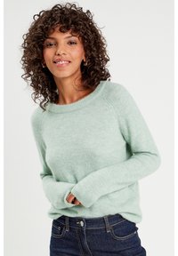 Suéter verde claro de tela suave, con cuello redondo, mangas largas y puños acanalados, combinado con jeans de mezclilla oscura.