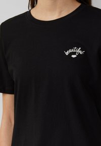 Zwart katoenen T-shirt met korte mouwen, voorzien van witte geborduurde tekst "beautiful" met een lipgraphic op de linkerkant van de borst.