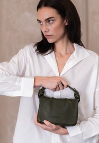 Olijfgroene leren handtas met een gestructureerd oppervlak, geknoopte handgreep en een rechthoekige vorm, vastgehouden door iemand in een wit shirt.