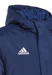 Marineblauwe jas met een capuchon, gemaakt van gladde stof, met een wit Adidas-logo aan de linkerkant. Gestructureerde accenten rond de kraag.
