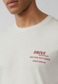 Witte katoenen T-shirt met een gestructureerde afwerking en een ronde hals, voorzien van rode bedrukte tekst: "DRIVE STUNT UNIT" en "MOTION PICTURES."