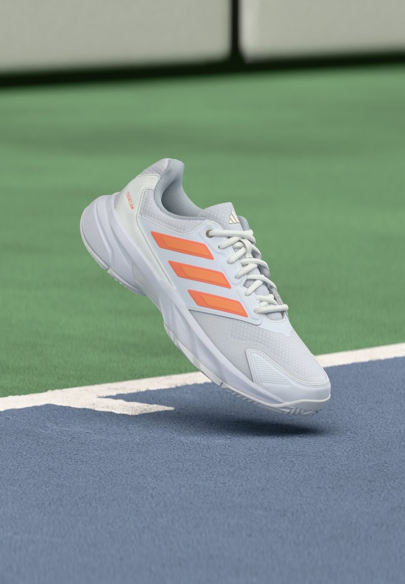 Γκρι αθλητικό παπούτσι με πορτοκαλί ρίγες, υφασμάτινο επάνω μέρος, κλείσι μο με κορδόνια και μαλακή σόλα, τοποθετημένο πάνω από μπλε επιφάνεια γηπέδου.