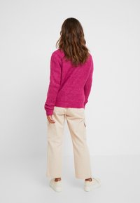 Femme aux longs cheveux bruns portant un pull rose vif, un pantalon large beige et des baskets blanches, debout de dos sur un fond uni.