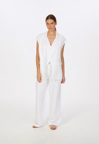 Weißes Leinenhemd mit aufgerollten Ärmeln, Knopfleiste und Seitentaschen, kombiniert mit lockeren weißen Hosen. Leichter, atmungsaktiver Stoff, minimalistisches Design.