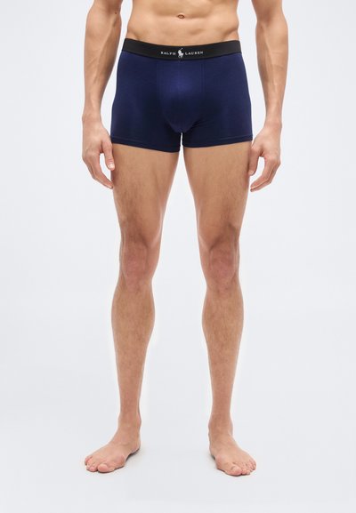 Άνδρας μοντέλο στέκεται ξυπόλυτος φορώντας μπλε Ralph Lauren ανδρικά μποξεράκια με μαύρη ζώνη στη μέση, πάνω σε απλό φόντο.