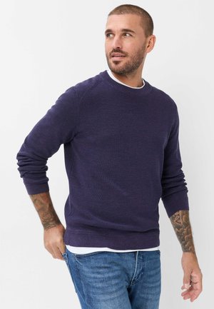 Man met kort haar en baard draagt een donkerblauwe trui en jeans, kijkt opzij met een neutrale uitdrukking, hand bij de zak.