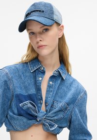 Denims krekls ar mezglu priekšā, ar gaiši un tumši zilu raibumu dizainu. Saskaņots ar zilu denim baseball cepuri.