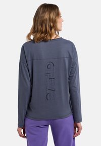 Grauer Langarm-Oberteil mit einem strukturierten "SAND"-Grafik auf dem Rücken. Hat einen gerundeten Ausschnitt und einen lockeren Schnitt, kombiniert mit lila Hosen.