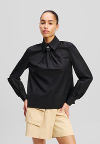 Schwarz langärmliges Hemd mit gerafftem Vorderteil, klassischem Kragen und silbernem Akzent. Kombiniert mit beigen Shorts mit Taschen.