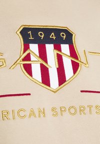 Tissu beige avec un logo en forme de bouclier comportant des rayures rouges et blanches, un sommet bleu foncé, une broderie dorée et l'année "1949". Le texte indique "AMERICAN SPORTS".