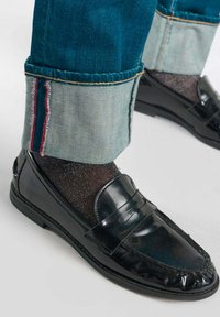 Mocassins en cuir verni noir associés à des chaussettes noires scintillantes. Jean en denim bleu avec des ourlets repliés mettant en valeur des coutures rouges.