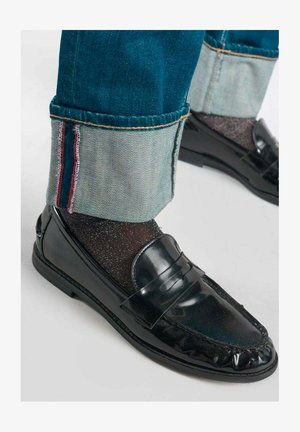 Schwarze Lackleder-slipper kombiniert mit glitzernden schwarzen Socken. Blaue Jeans mit umgeschlagenen Bündchen, die eine rote Stichdetail zeigen.