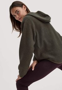 Olivgr�üner Fleece-Hoodie mit strukturierter Oberfläche, lockerer Passform und vorderer Kängurutasche, getragen über dunkellila Leggings.