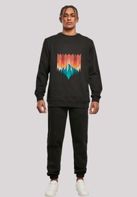Schwarzer Sweatshirt mit einem bunten geometrischen Grafik von Bergen und Wäldern. Kombiniert mit schwarzen Jogginghosen und weißen Sneakern.
