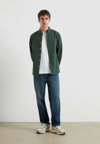 Donkergroene knoopshirt over een wit T-shirt, gecombineerd met blauwe jeans en grijzenn sneakers. Het shirt heeft een klein logo op de linkerborst.