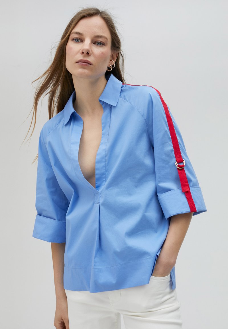 Camicia azzurro chiaro con scollatura a V profonda, maniche arrotolate e strisce rosse lungo le braccia. Tessuto morbido con vestibilità comoda.