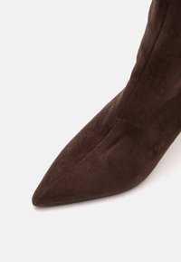 Gros plan d'une botte haute en daim marron à bout pointu sur un fond clair uni.