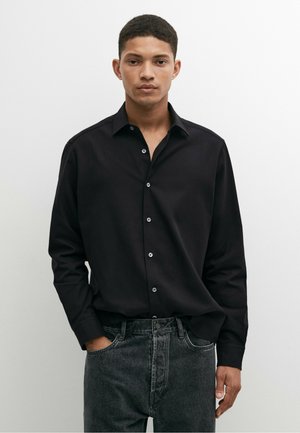 Giovane uomo con capelli corti che indossa una camicia nera con bottoni e jeans scuri, in piedi con una mano in tasca davanti a uno sfondo bianco uniforme.