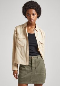 Cămașă bej cu butoane, având două buzunare pe piept, purtată peste un top negru fără mâneci, asortată cu o fustă verde măsliniu, ce dispune de buzunare frontale și un buton la talie.