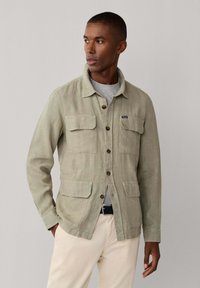 Uomo che indossa una giacca beige con bottoni e quattro tasche sopra una camicia grigia e pantaloni color crema, che guarda lateralmente contro uno sfondo semplice.