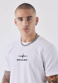 T-shirt bianca a maniche corte con logo nero "PXP" e design a stella. Il modello indossa una collana di catena d'argento spessa e ha tatuaggi visibili.