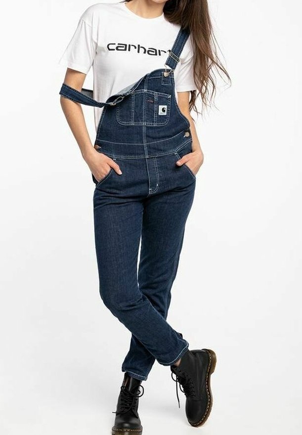 Femme portant une salopette en denim bleu foncé avec une bretelle détendue, un t-shirt blanc Carhartt et des bottes noires à lacets, debout les mains dans les poches.