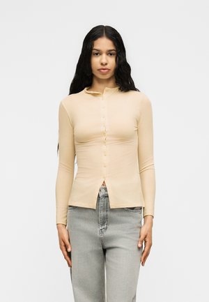 Donna con capelli lunghi e scuri che indossa una maglia beige a maniche lunghe con bottoni e jeans grigio chiaro, in piedi davanti a uno sfondo chiaro e uniforme.