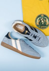Paire de baskets en daim bleu clair avec des rayures blanches et des semelles marron, placées à côté d'une chemise jaune avec un logo circulaire vert.