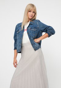 Blond kvinde iført en blå denimjakke over en hvid top og en lang, plisseret, skinnende sølvskørt, der poserer mod en grå baggrund.